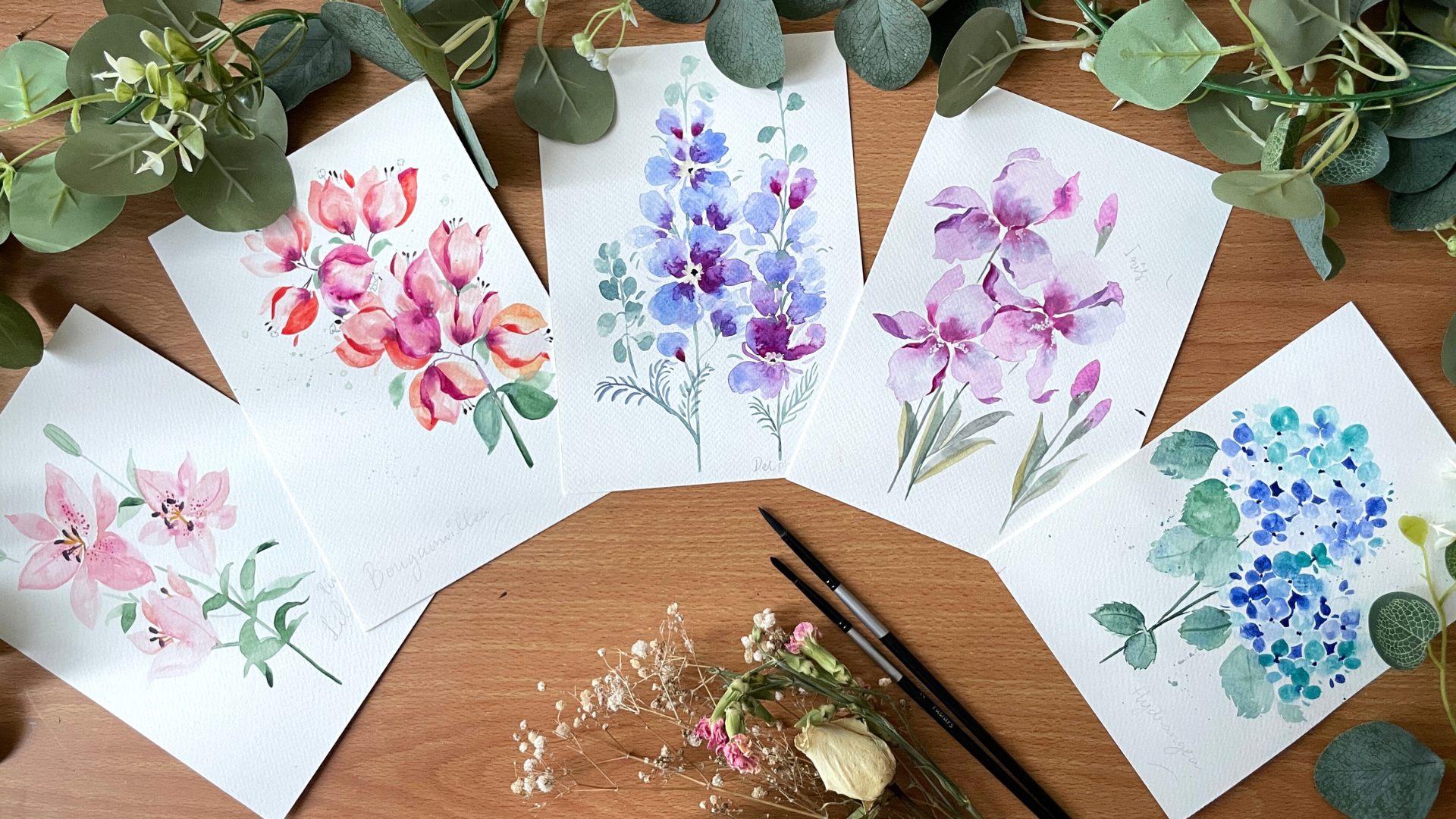 Watercolor paintings of flowers on white paper with green leaves and brushes on a wooden surface. Femvisionary painting